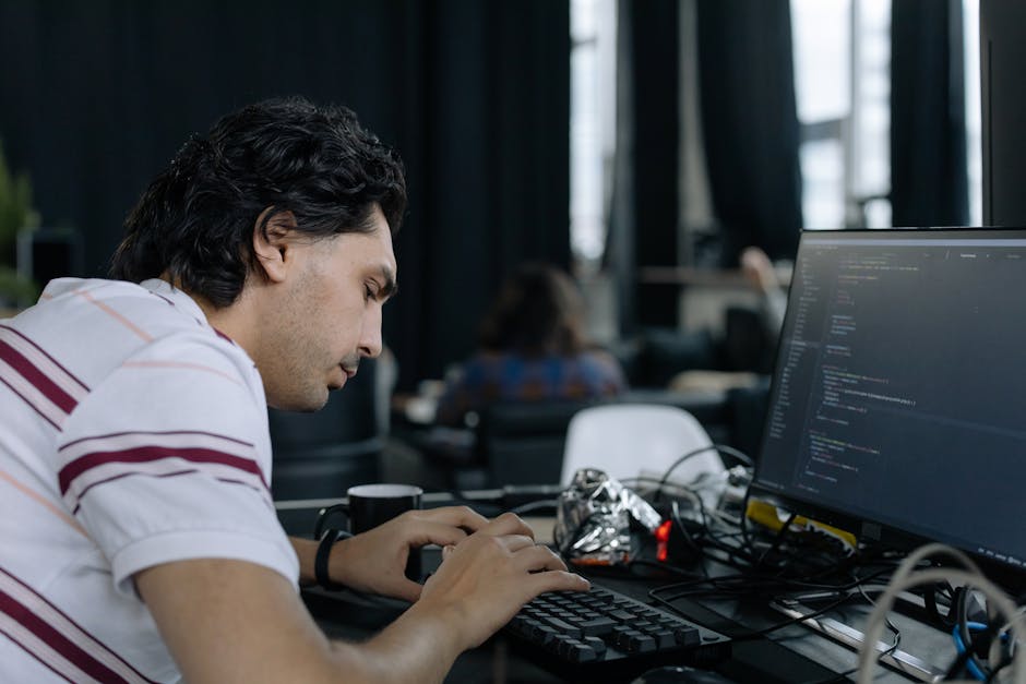 A young man with dark, wavy hair and light skin is working at a desk in a modern office environment, focusing intently on a computer monitor displaying lines of code. He is wearing a white shirt with 