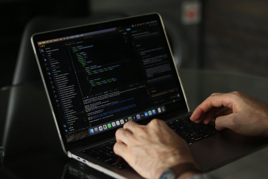 A young man with dark, wavy hair and light skin is working at a desk in a modern office environment, focusing intently on a computer monitor displaying lines of code. He is wearing a white shirt with 