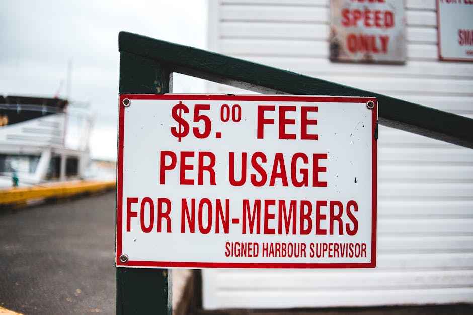 A close-up view of a white rectangular sign with bold red lettering, displaying a fee of $5.00 per usage for non-members, signed by the Harbour Supervisor, mounted on a dark green wooden post. In the